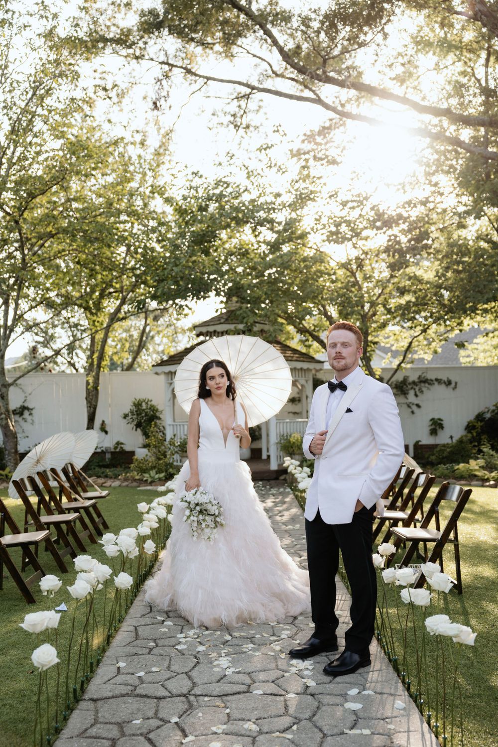 Wedding couple poses on stone path lined with white flowers and garden gazebo backdrop.