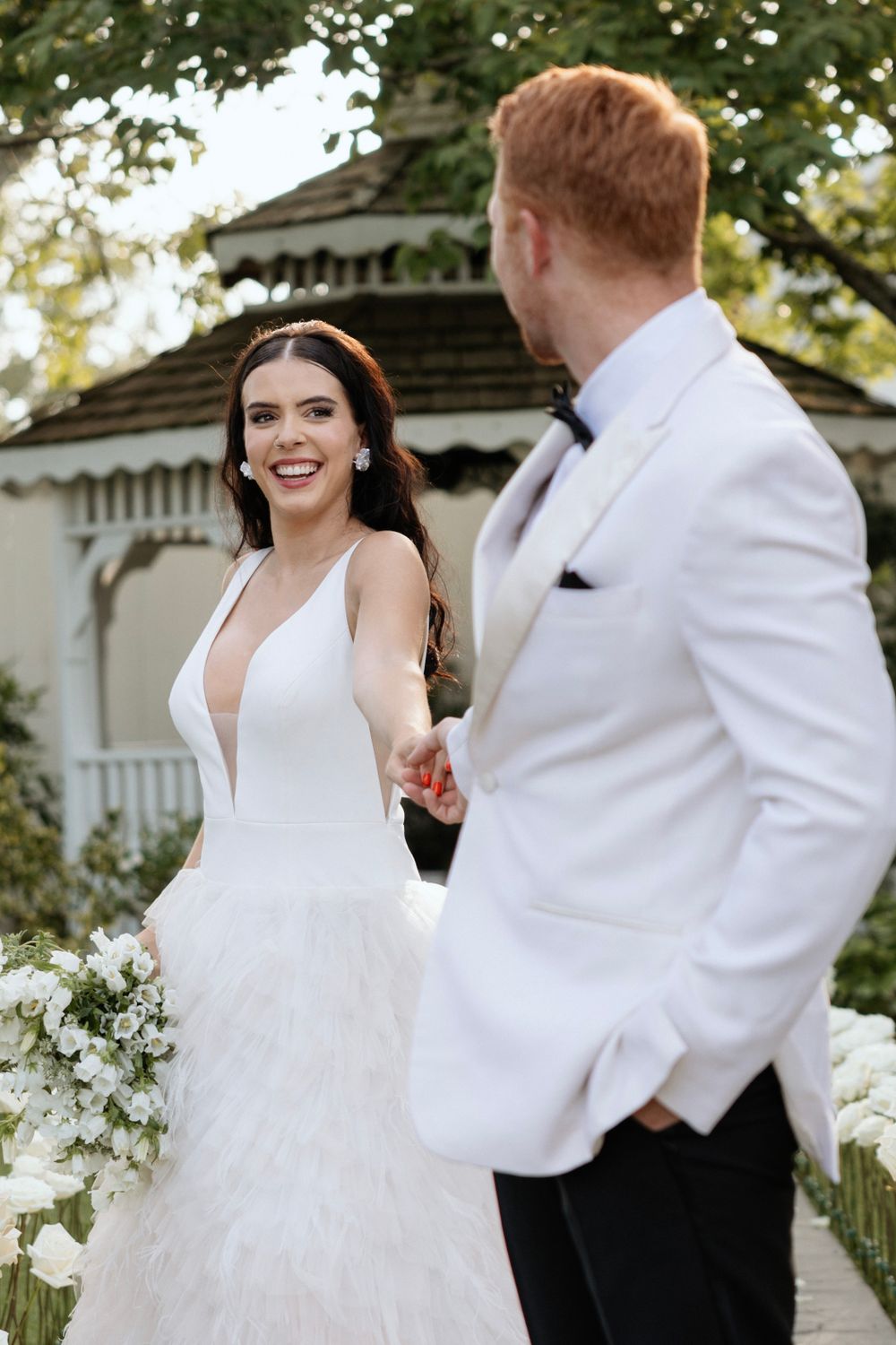 Newlyweds share a joyful moment in front of garden gazebo surrounded by white floral decorations.