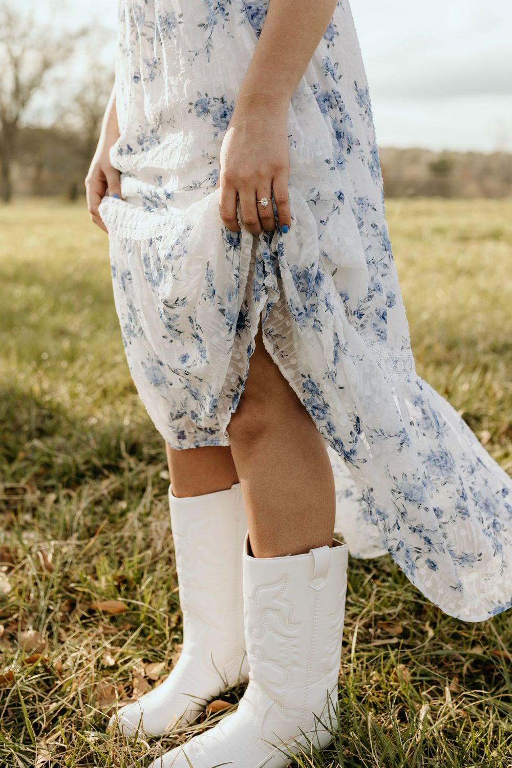 A close-up of white cowboy boots and a floral dress hem in a rustic outdoor setting.