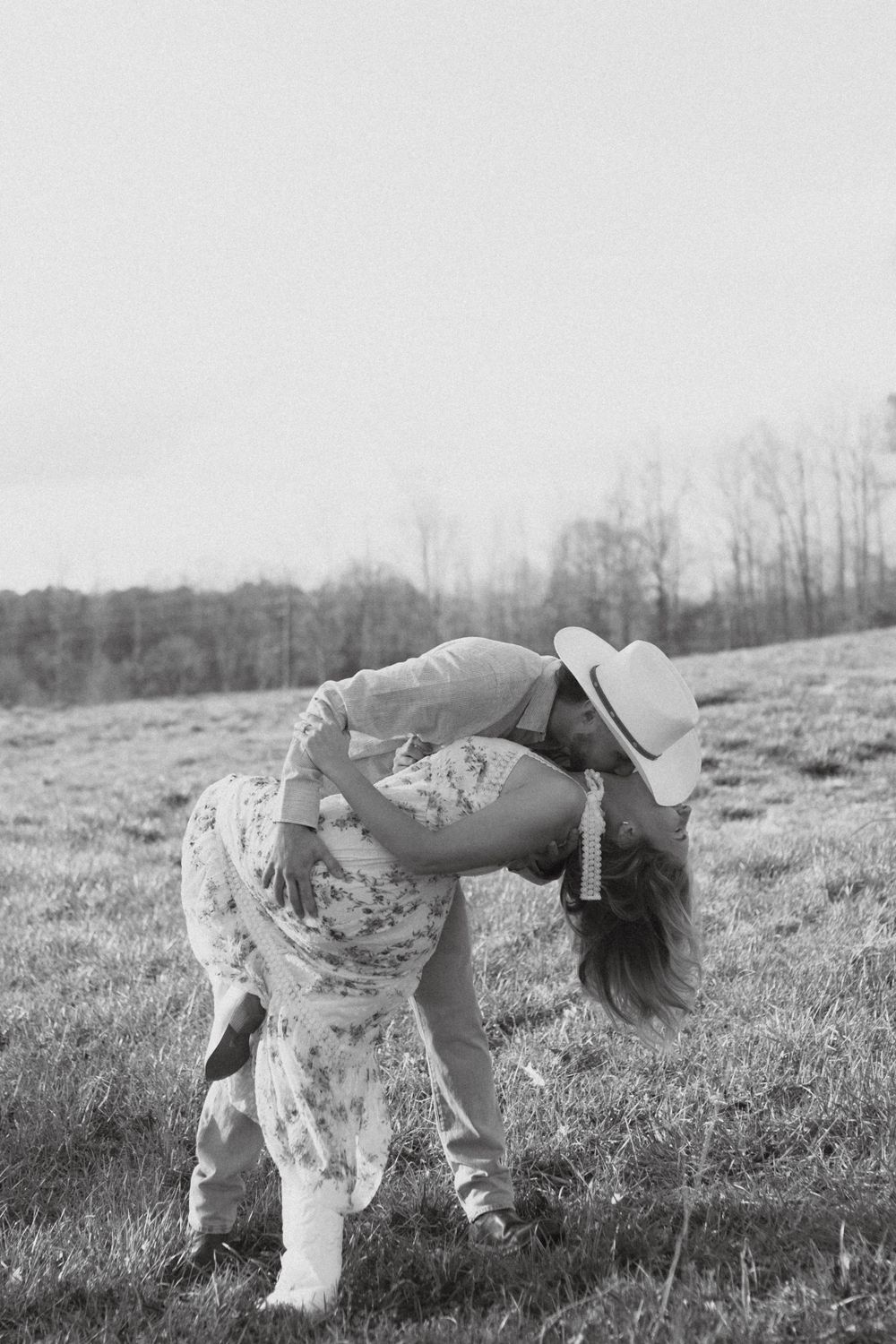 A romantic couple in a countryside field, with a floral dress and cowboy hat creating a playful moment.