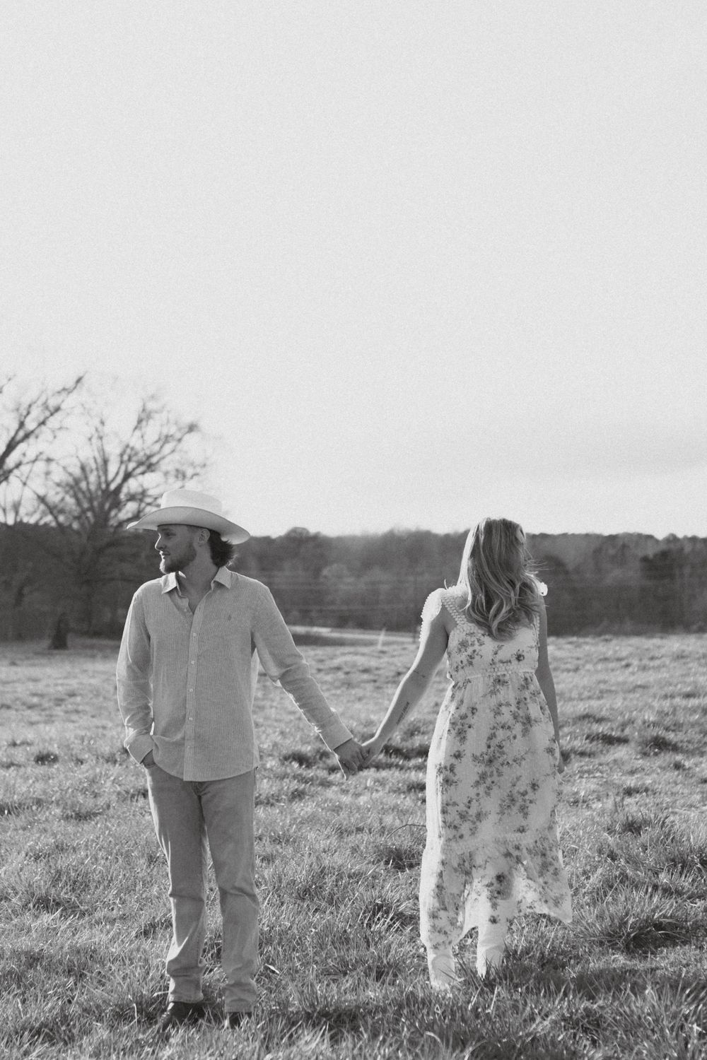 A couple walking hand in hand through a pastoral field in western-inspired outfits.