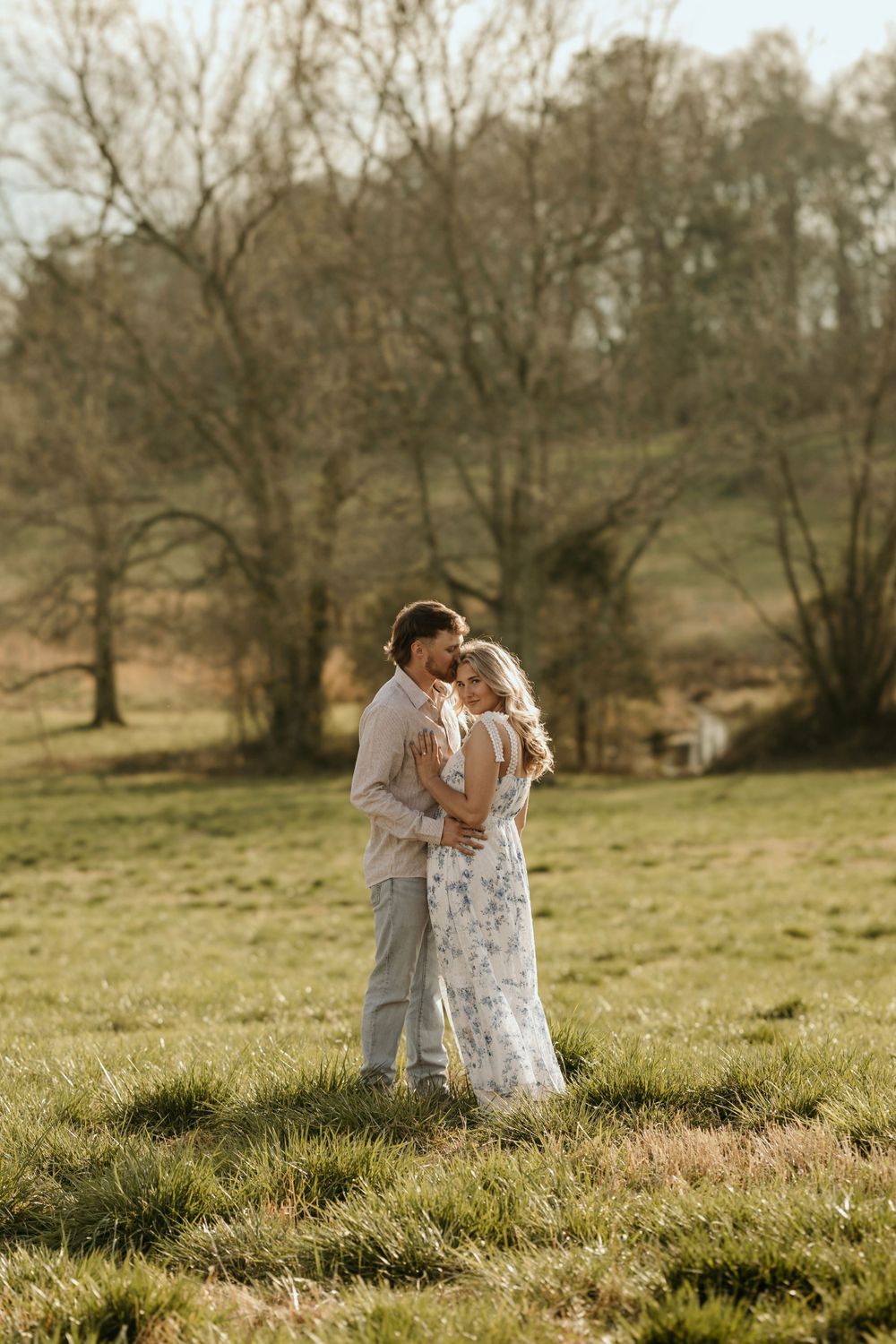 A tender embrace between newlyweds in a sunlit meadow with bare trees in the background.