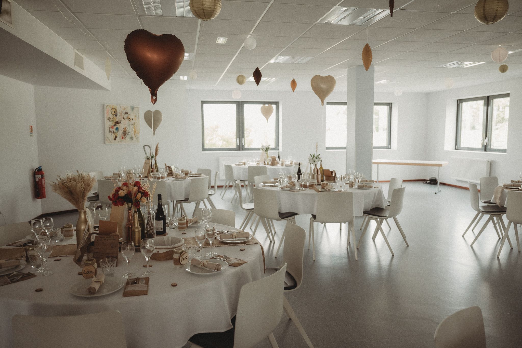 White modern reception hall decorated with heart balloons and round tables for a wedding celebration.