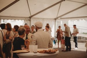 Reception guests mingling near food tables under a white tent at a casual celebration.
