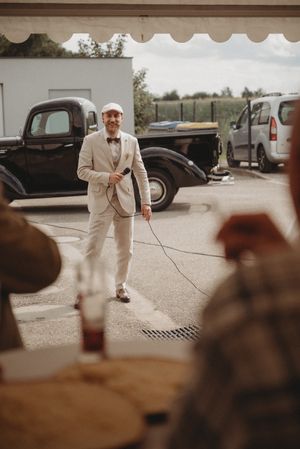 Person in light suit stands near vintage car at outdoor event.