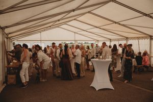 Large group of wedding guests mingling under white reception tent with cocktail tables.