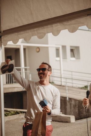 An entertainer in sunglasses and white sweater speaks into a microphone at an outdoor event.
