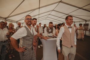 Wedding guests in formal attire gathered around tables under white tent reception.
