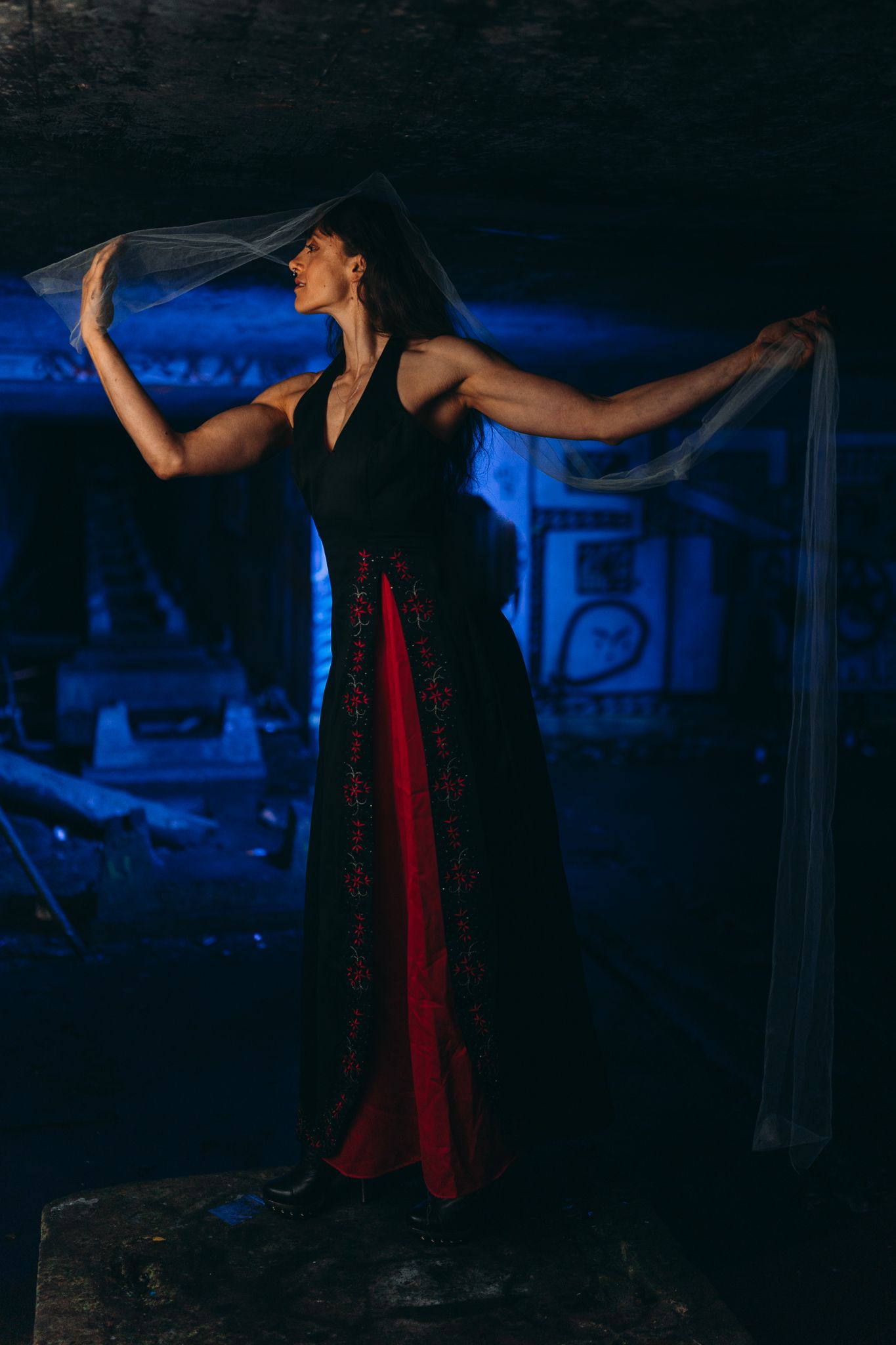 A dancer in a black dress and red sash performs an expressive movement sequence in a dark blue-lit studio space.