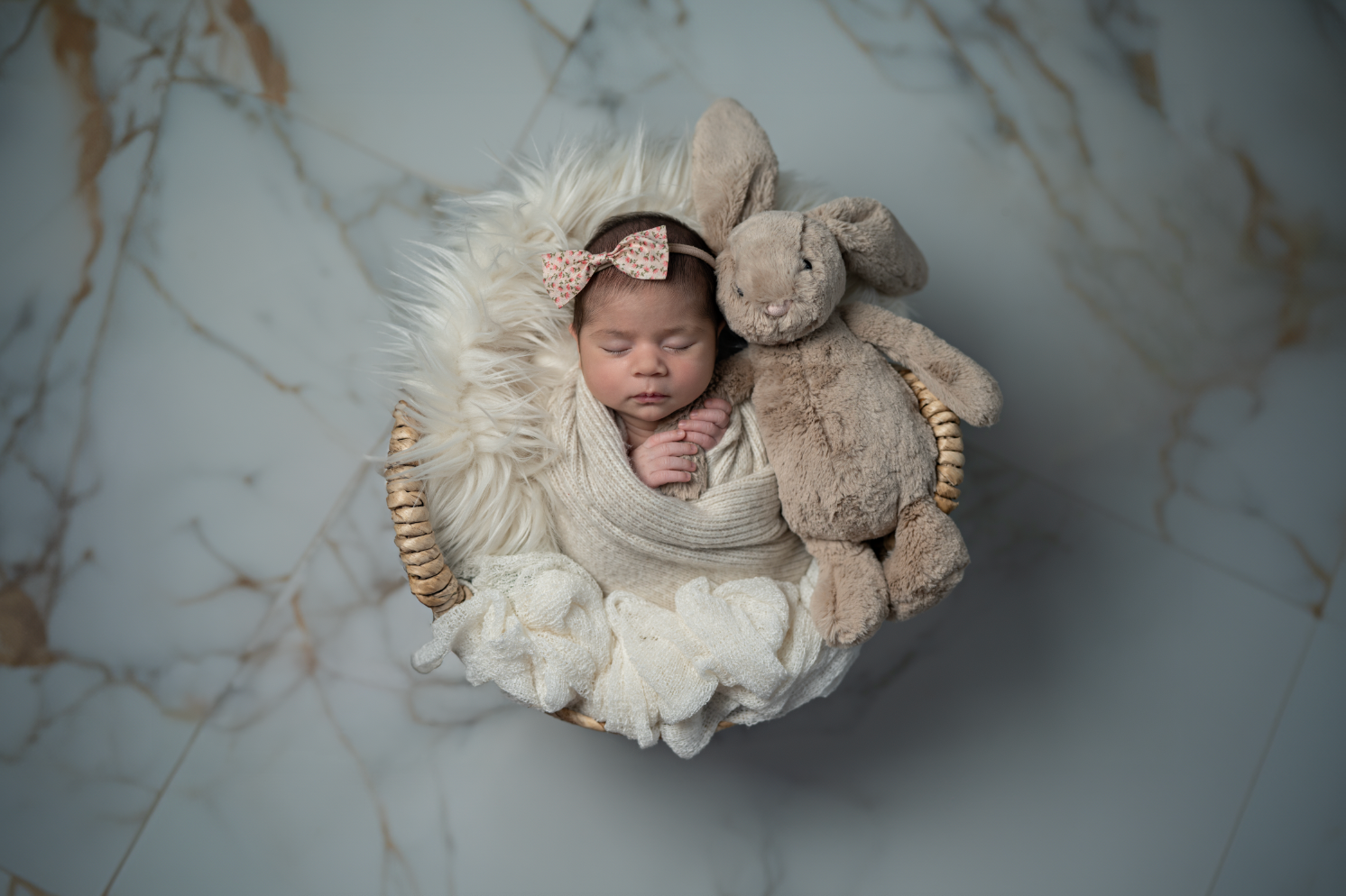 Newborn baby sleeping in a basket with a plush bunny toy, wrapped in cream fabric with a bow headband. Embracing Life’s Beauty
