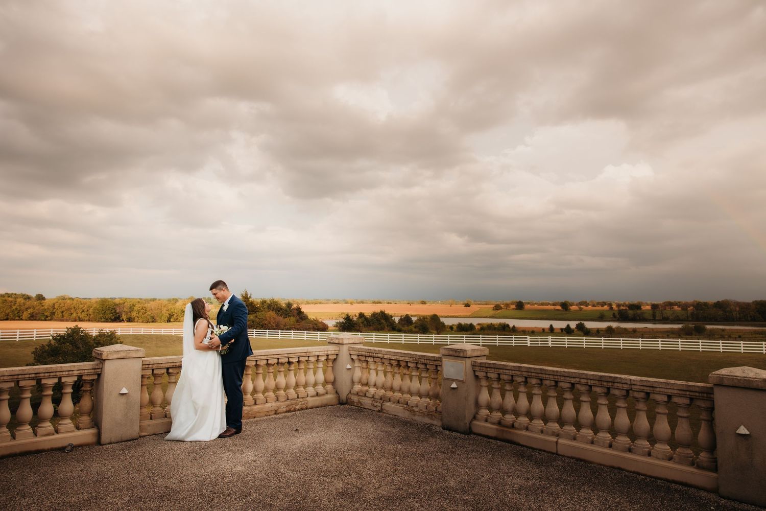 A wedding couple embracing on a stone balcony under dramatic cloudy skies.