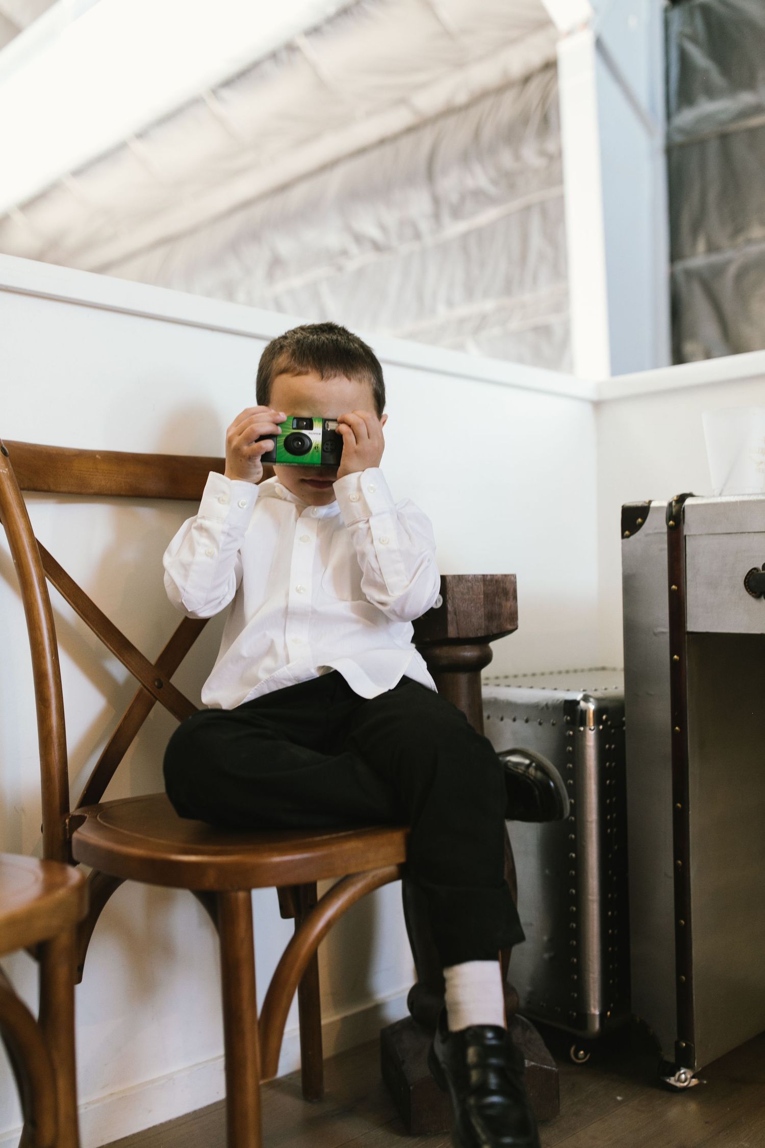 Small child in formal attire holds up a vintage camera while sitting on wooden chair.