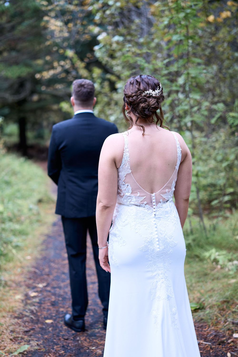 Back view of bride in lace-detailed dress walking forest path with groom.