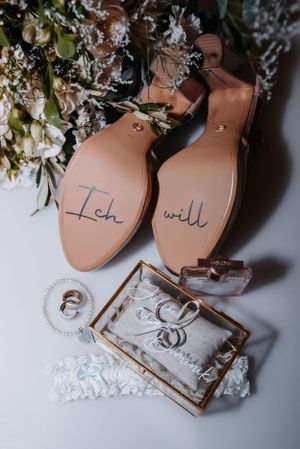 Wedding details flatlay with 'Ich will' written on shoe soles, rings and floral accents.