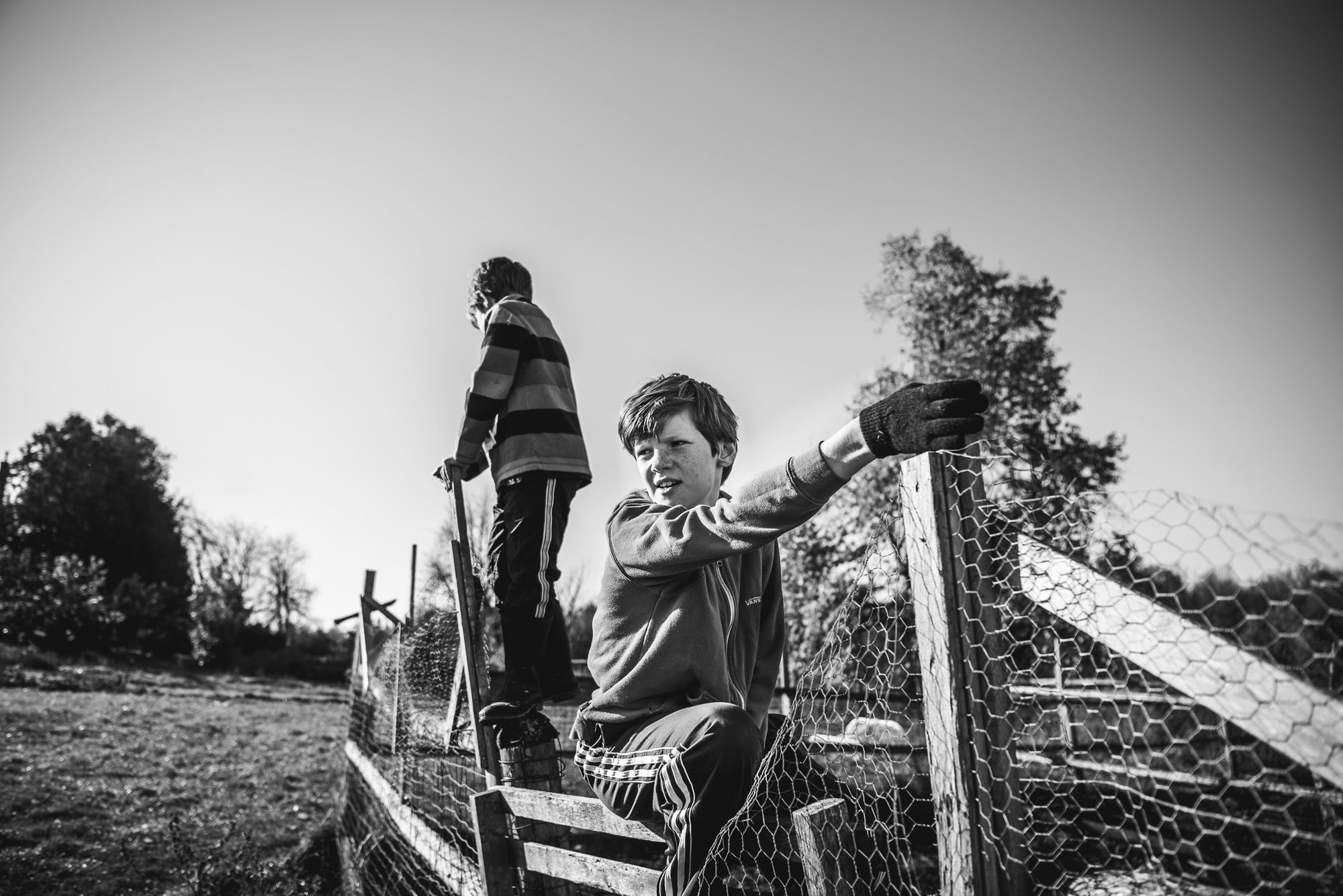 BW photo of a young man and his younger brother sitting on a wooden farm fence.