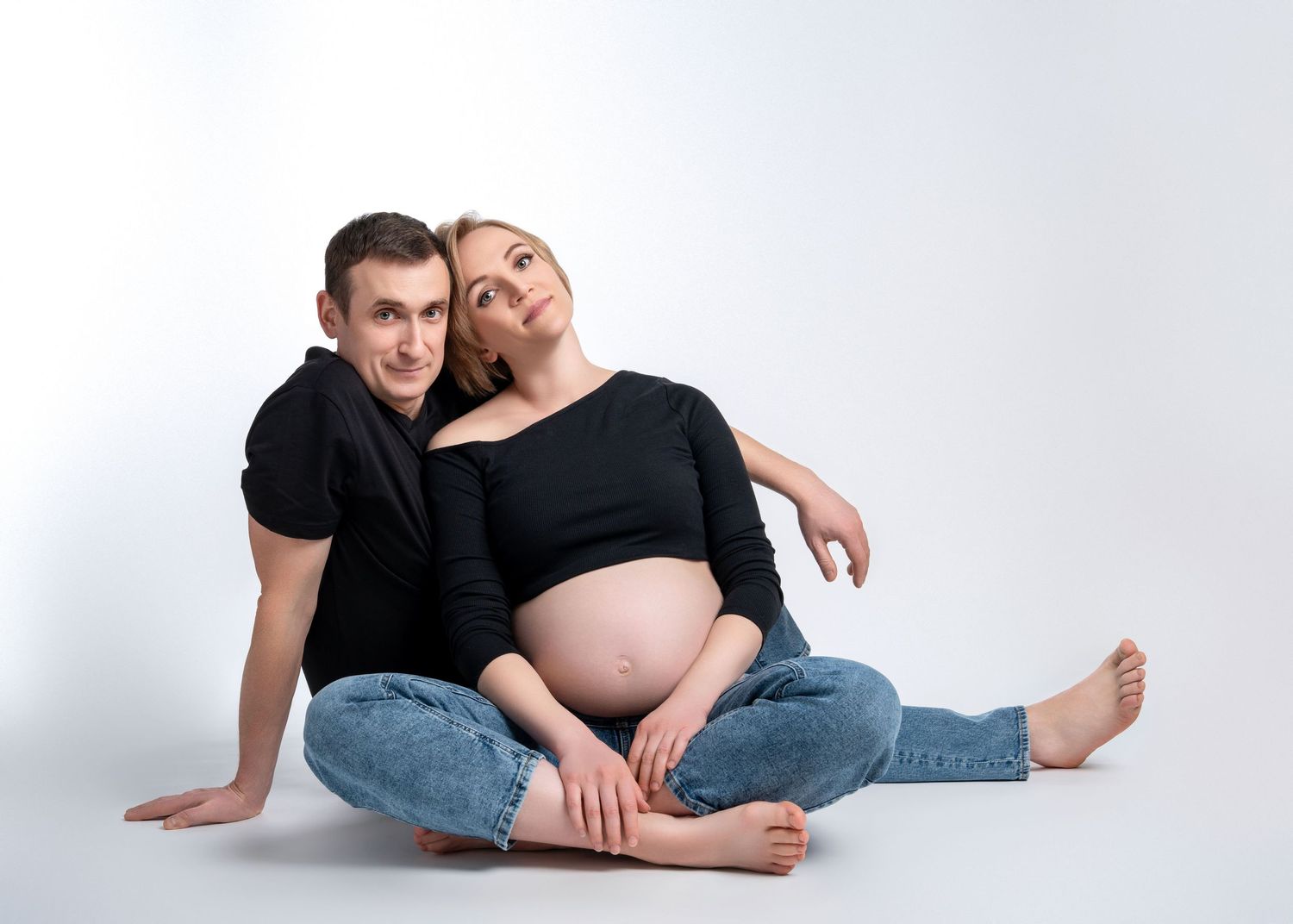 a pregnant woman sits and leans against her partner while wearing matching black tops and jeans