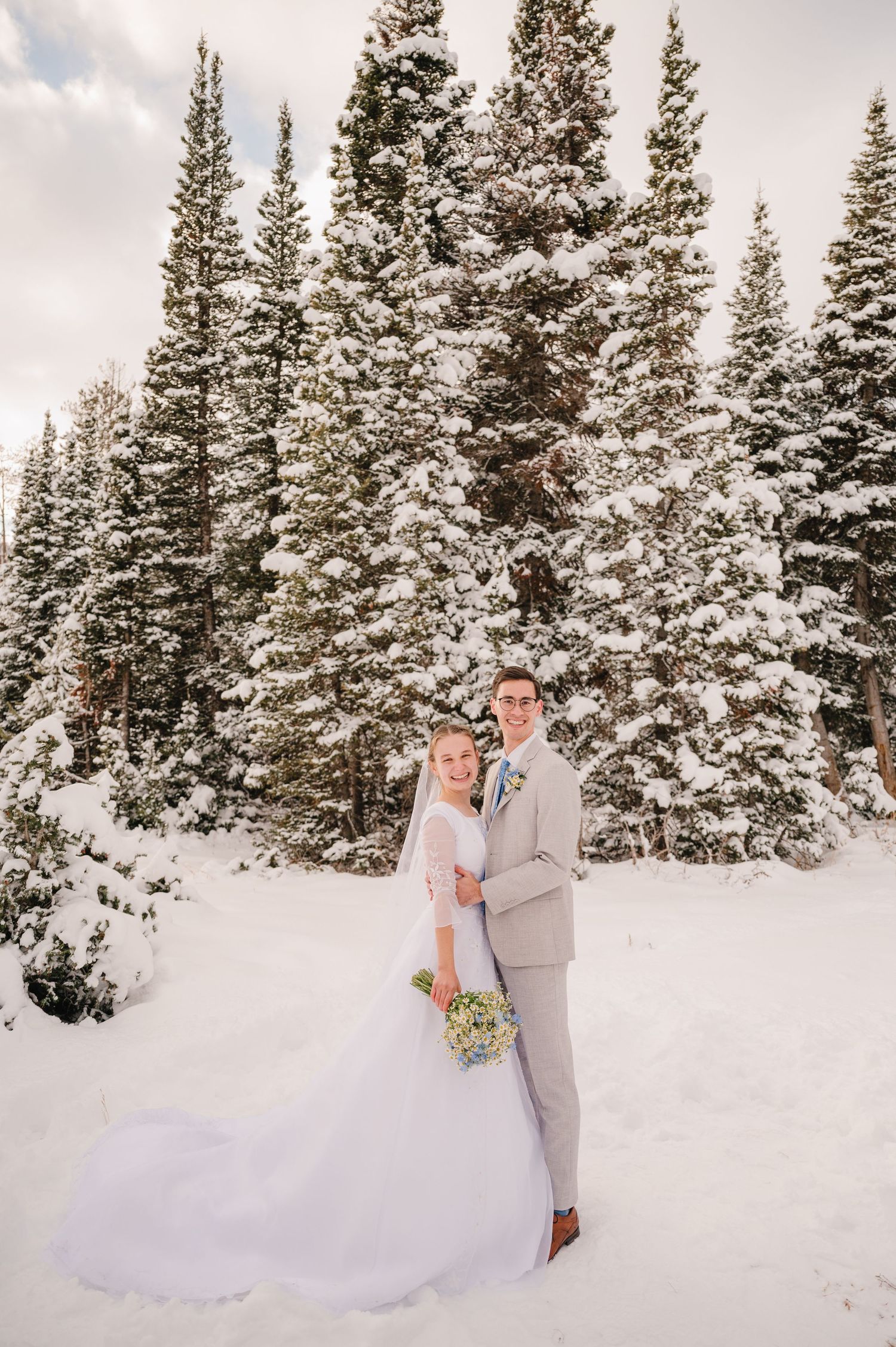 A newlywed couple stands before tall snow-covered pine trees in winter.