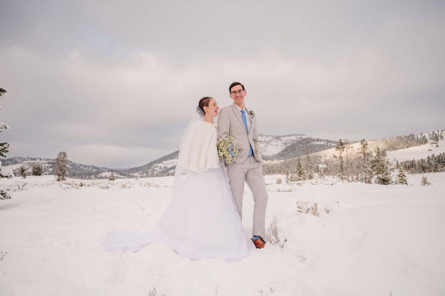 A bride and groom pose together in a snowy mountain landscape.