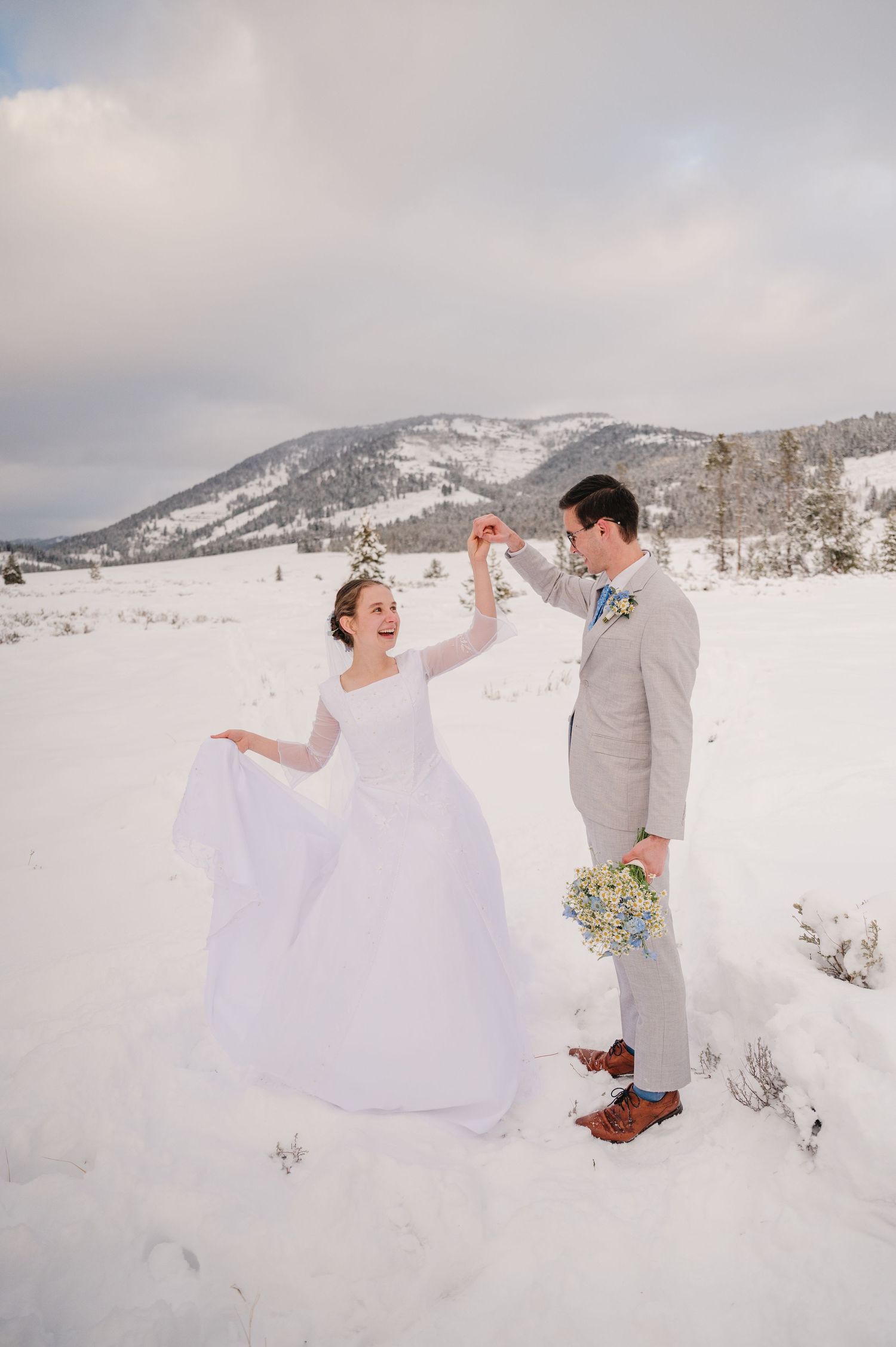 A joyful wedding couple plays in the snow with mountains in the background.