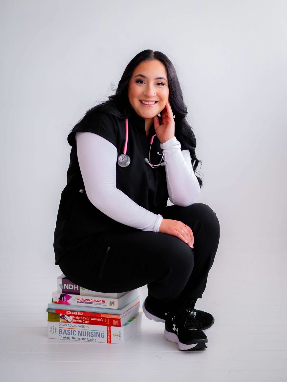 Smiling nurse in black scrubs and stethoscope posing with nursing textbooks in a professional photo.