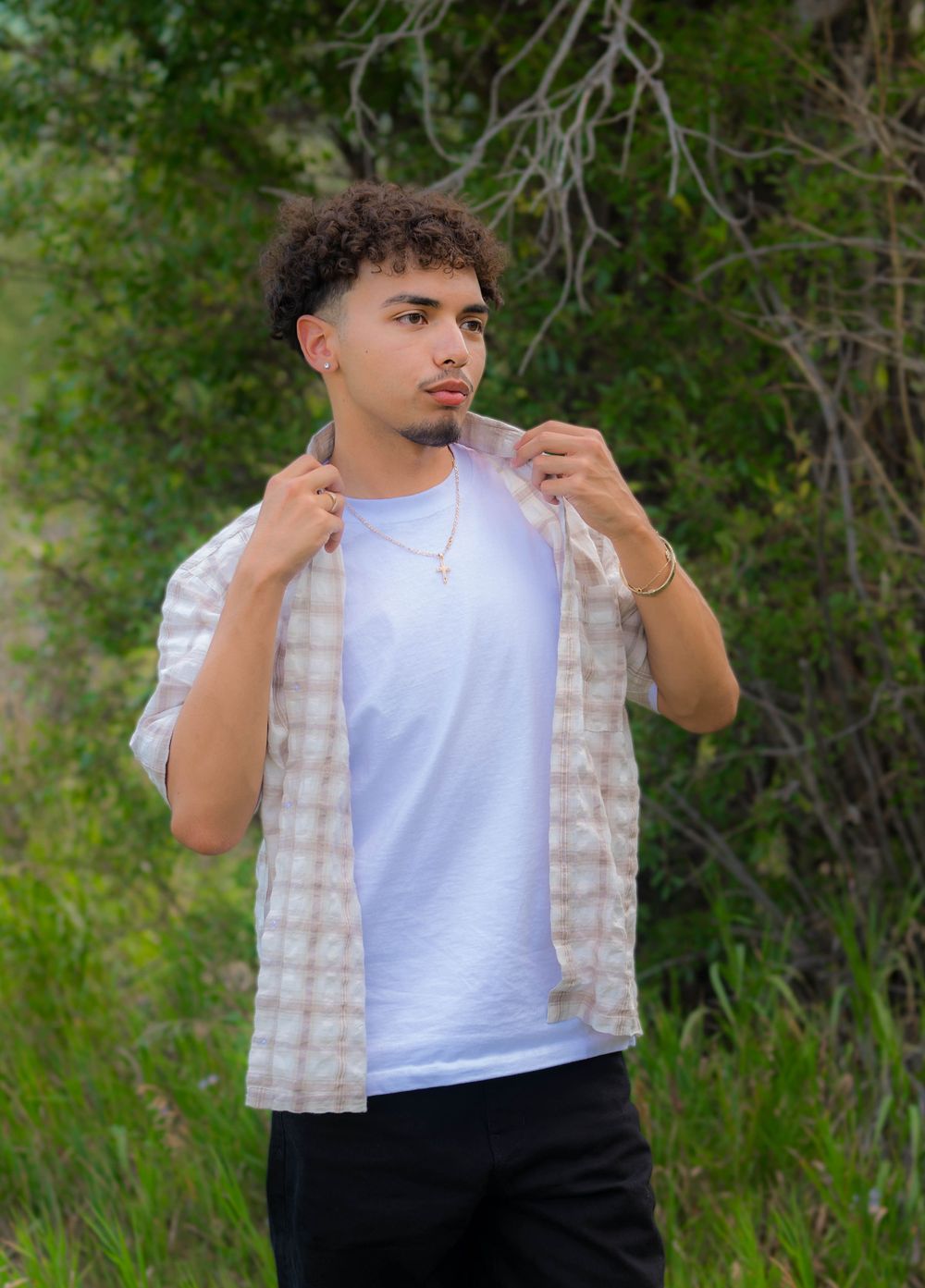 Young man in white tee and plaid shirt poses outdoors among lush green trees and shrubs.