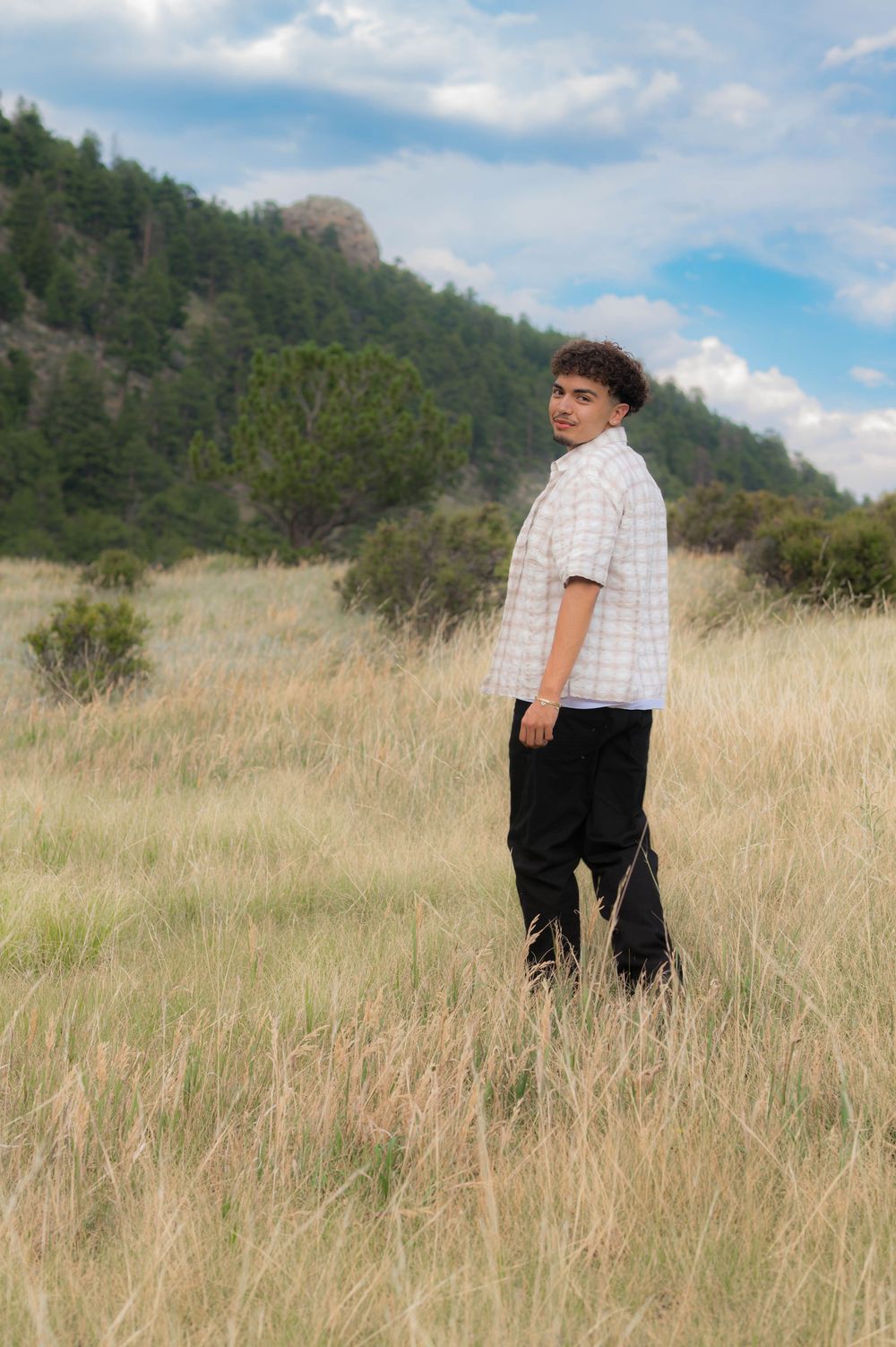 Young man in plaid shirt and black pants standing in tall grass field with mountain and trees in background.