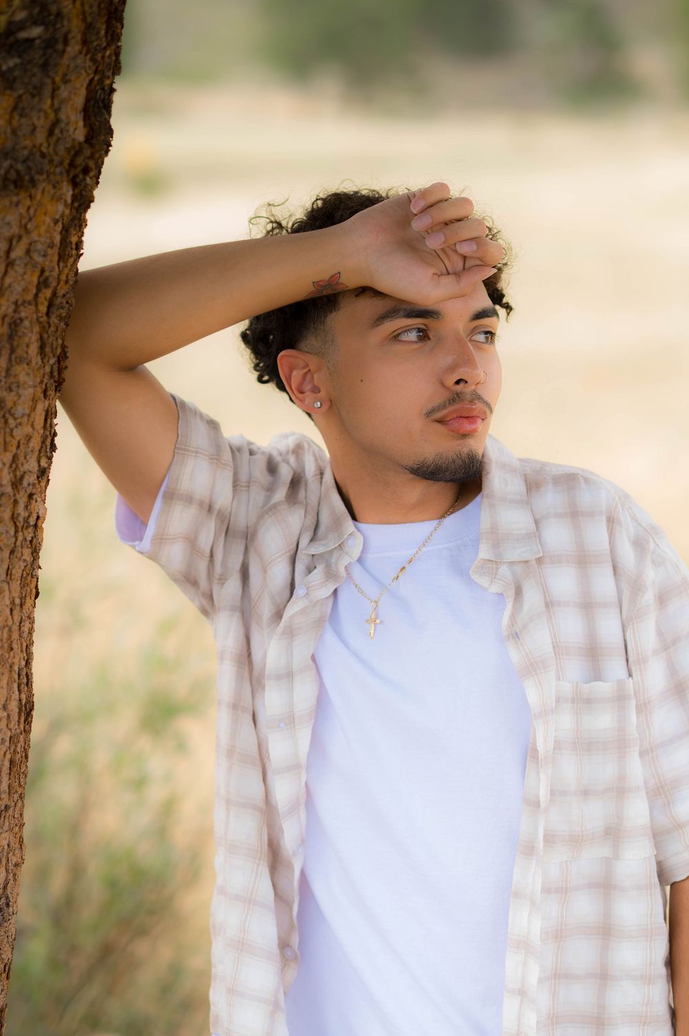 Young man in plaid shirt leaning against tree outdoors, hand on forehead, wearing cross necklace.