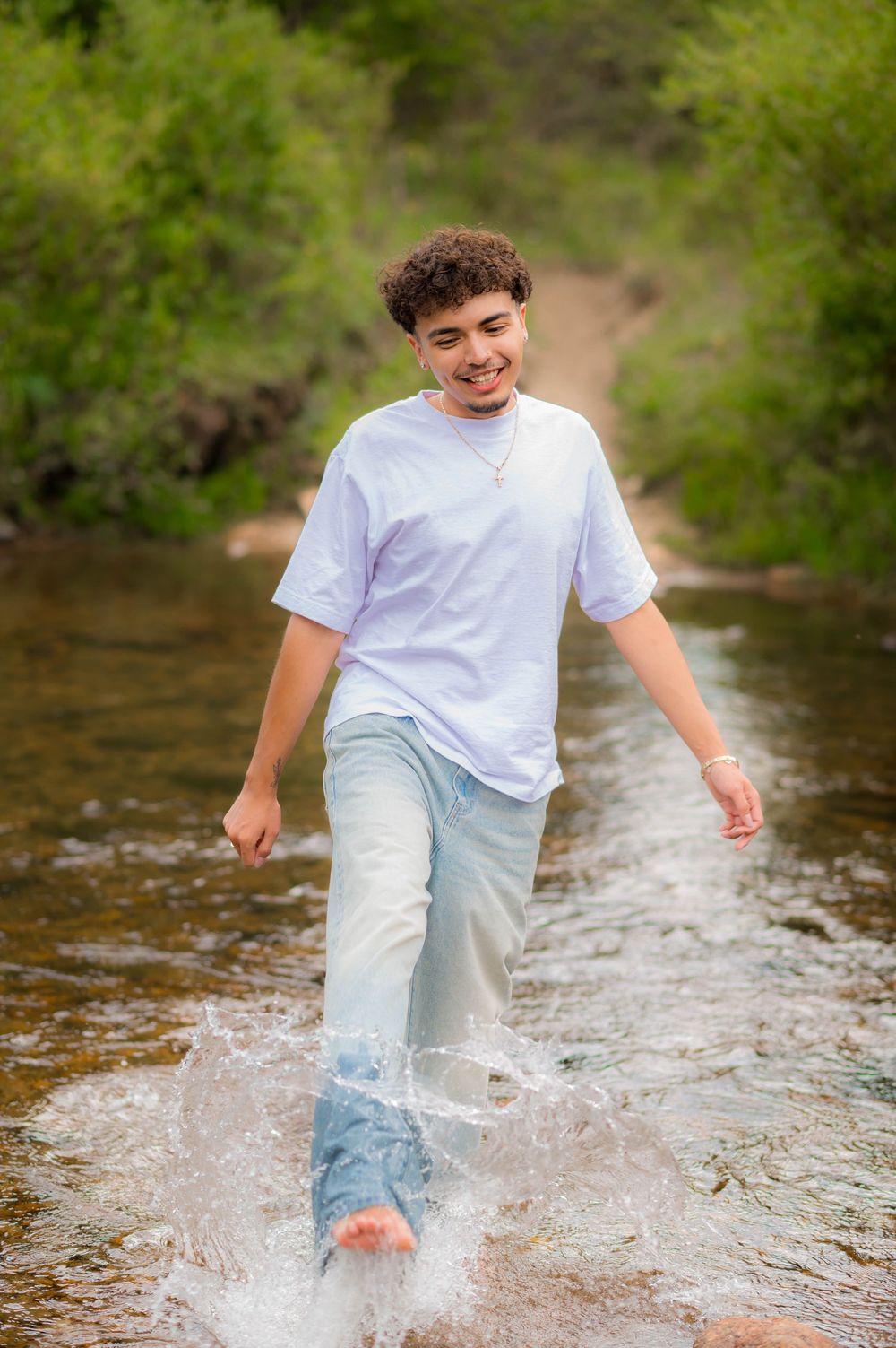 Smiling teen wading barefoot through a shallow creek, wearing a white t-shirt and light jeans, surrounded by green trees.