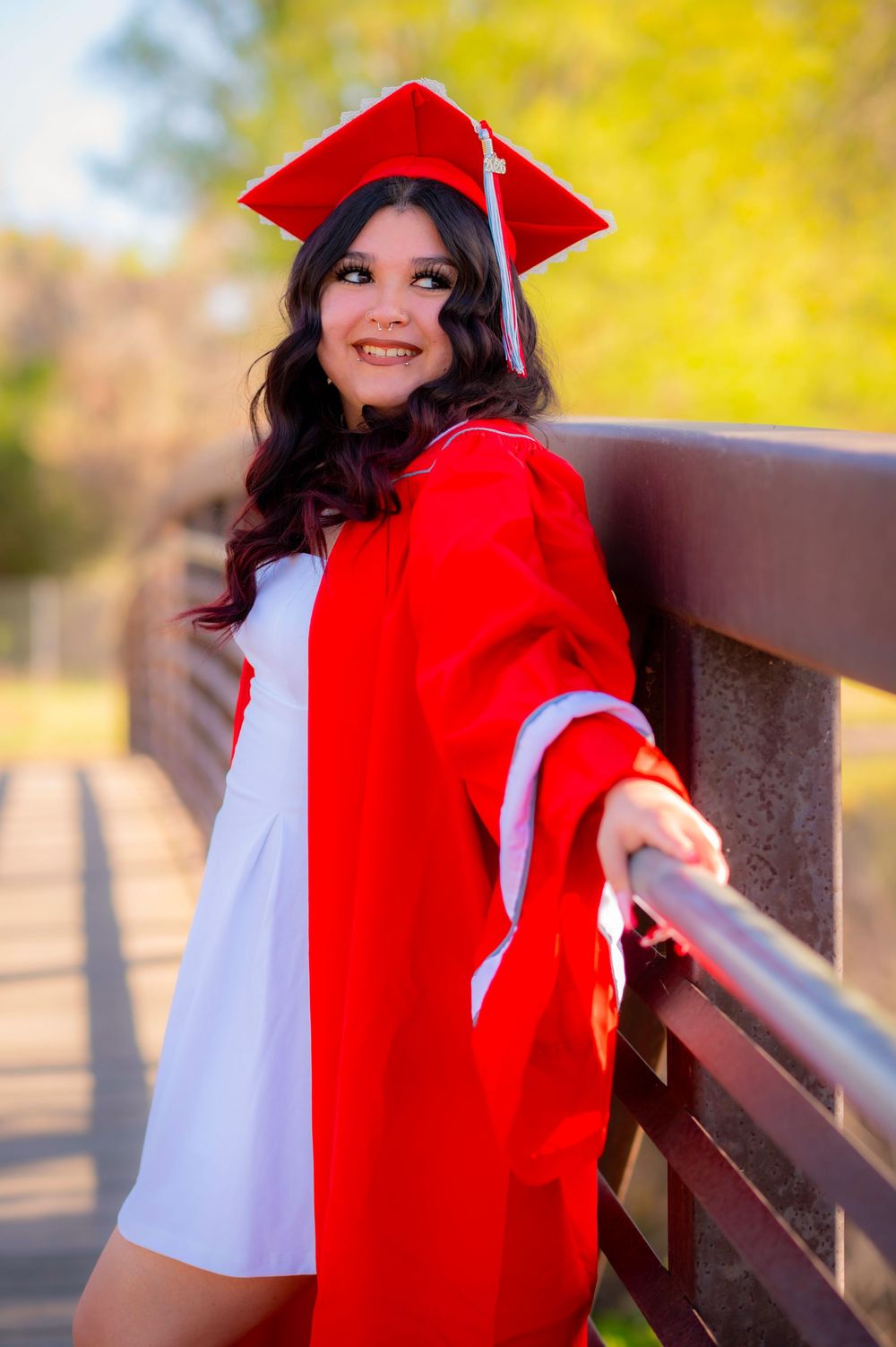 Smiling young woman in red graduation cap and gown posing on a bridge outdoors on a sunny day.