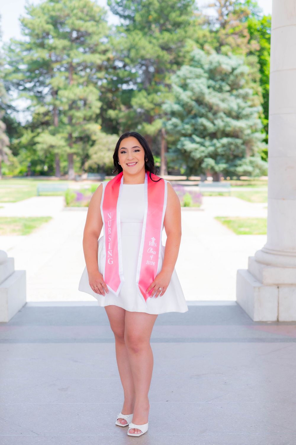Smiling graduate in white dress and pink sash poses between white columns with green trees in background.