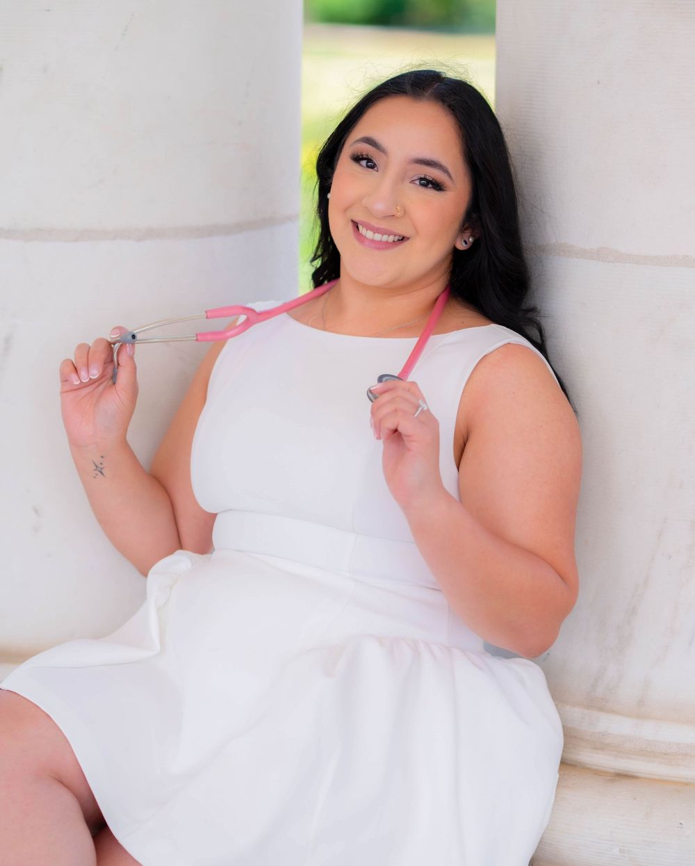 Young woman in white dress holding pink ribbon, smiling beside white columns outdoors.