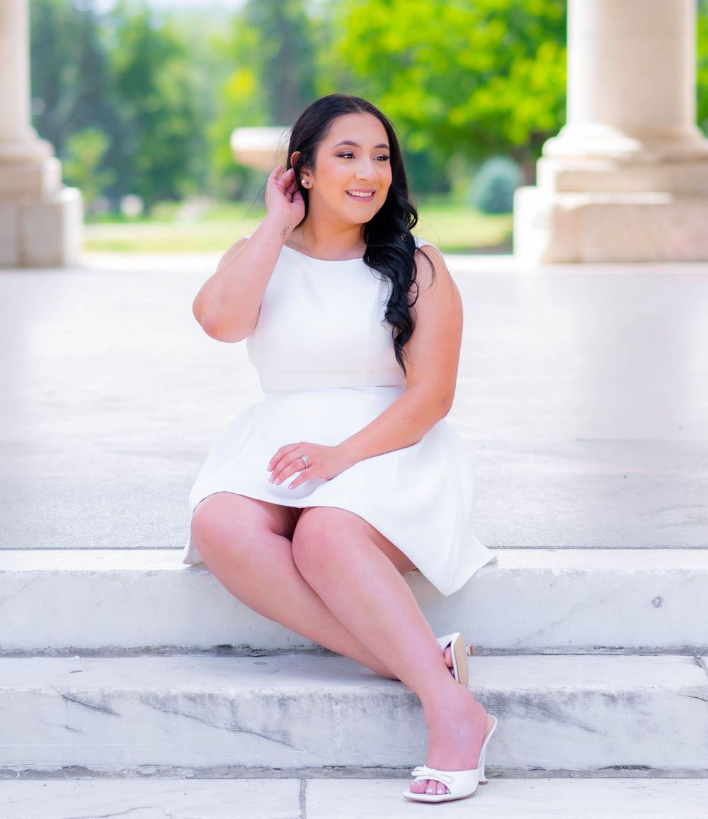 Young woman in white dress and heels sitting gracefully on marble steps outdoors near elegant columns.