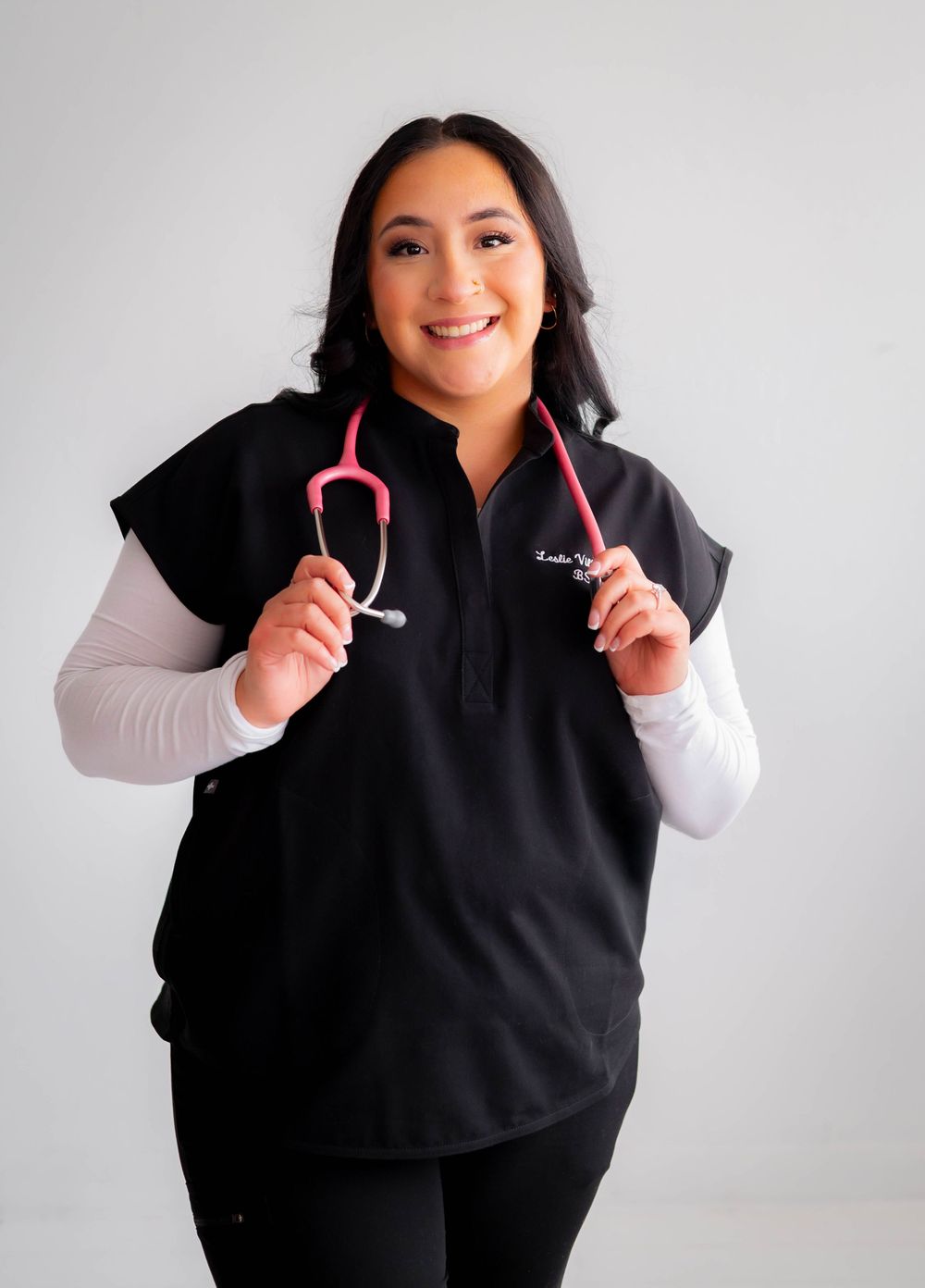 Smiling female healthcare professional in black scrubs vest holding a pink stethoscope against white background.