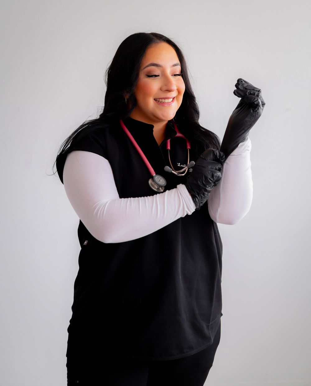 Smiling female healthcare worker in black scrubs and gloves with red stethoscope posing confidently.