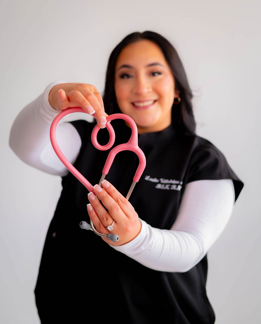 A smiling nurse in black scrubs holds a pink stethoscope shaped into a heart toward the camera.
