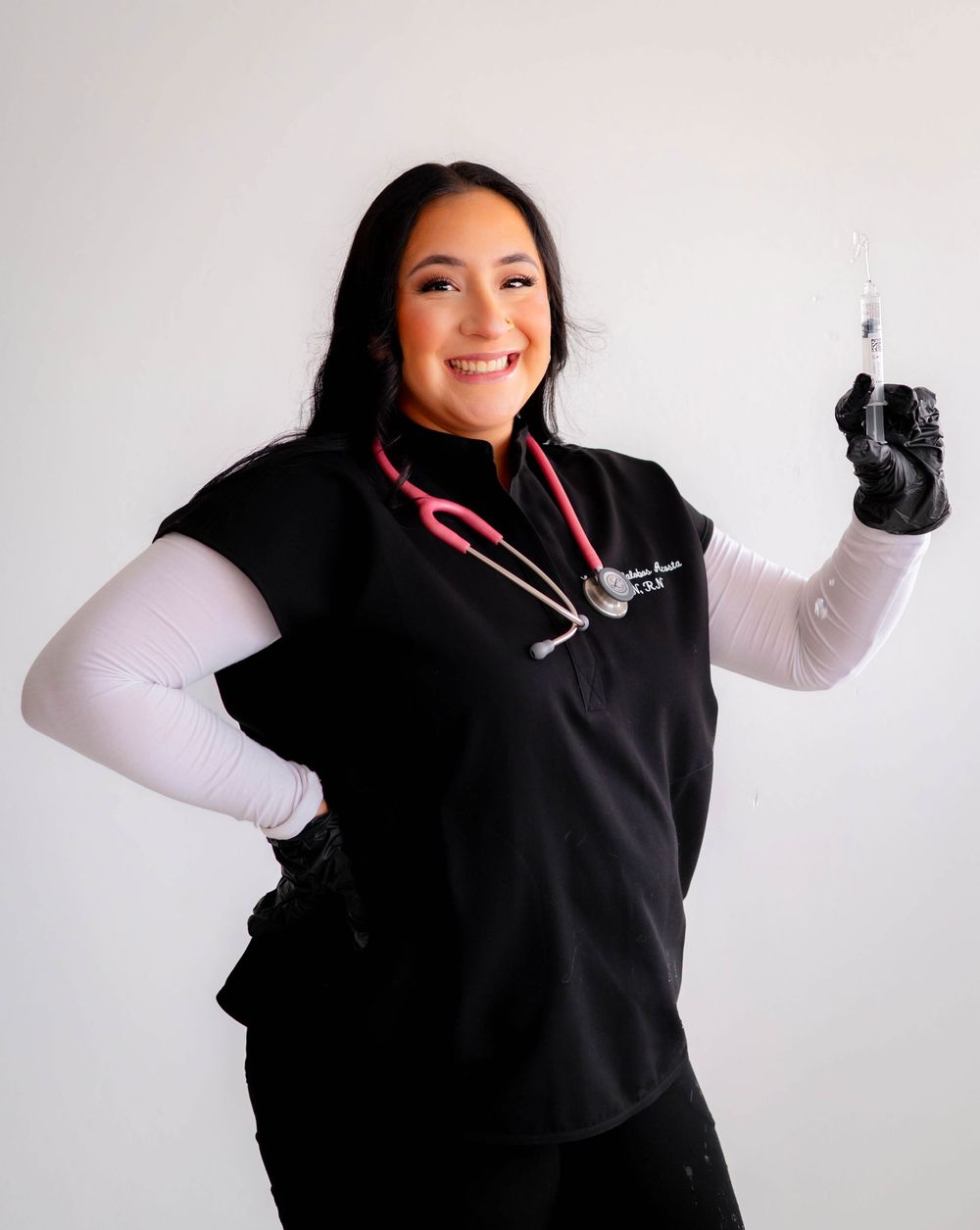 Smiling nurse in black scrubs with pink stethoscope holding a syringe, wearing black gloves and white undershirt.
