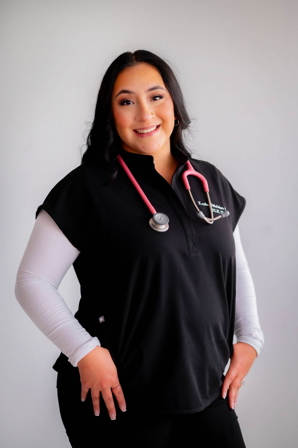Smiling female healthcare professional in black scrub vest with pink stethoscope posing against gray background.
