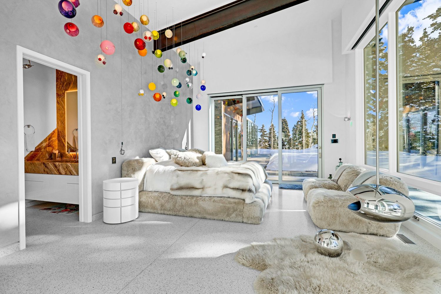 Contemporary white bedroom with colorful ceiling installation, floor-to-ceiling windows, and snowy forest view. Captured by Slate Media (bes