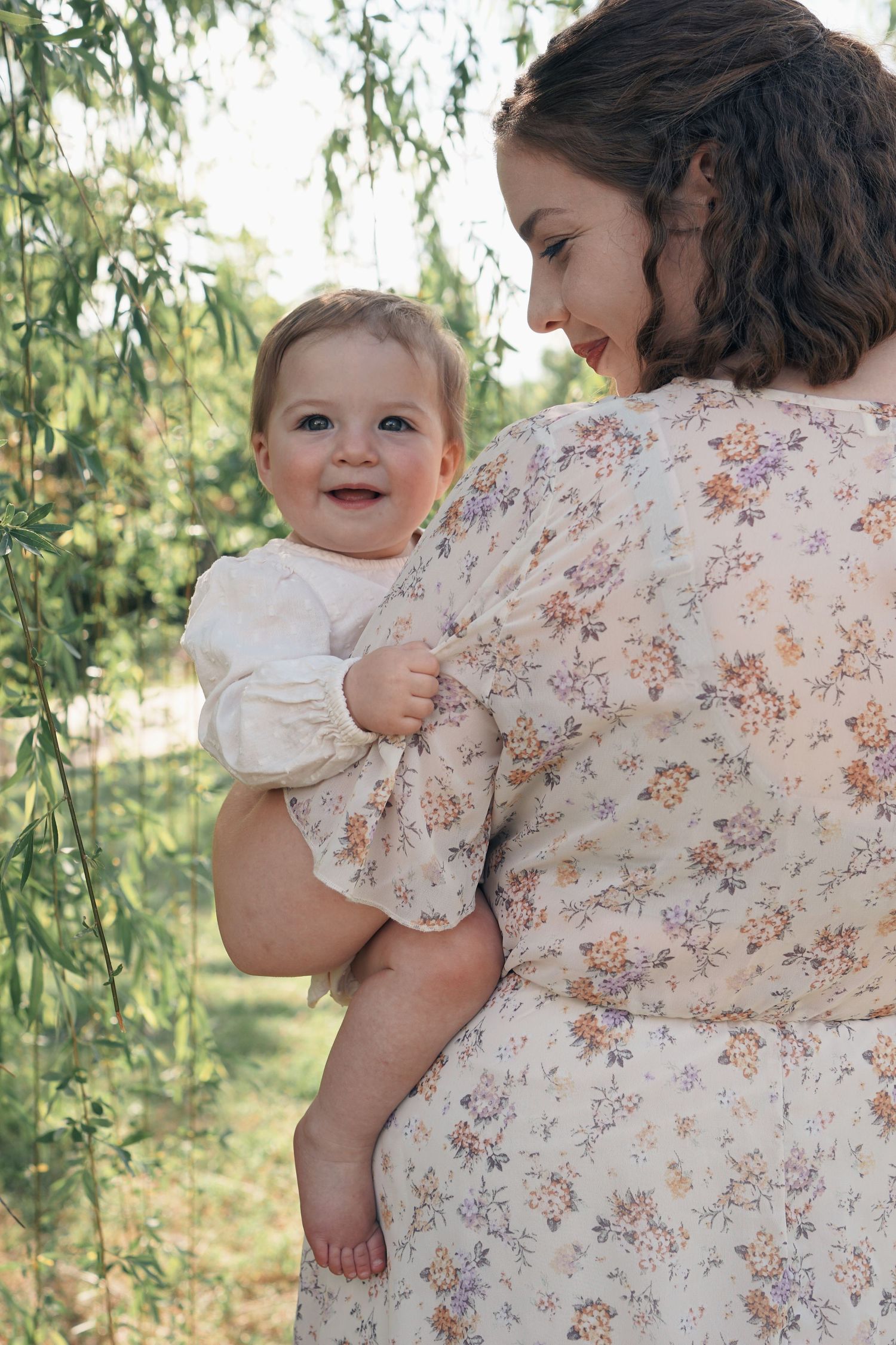 Parent and baby share tender moment among willow branches in springtime setting.