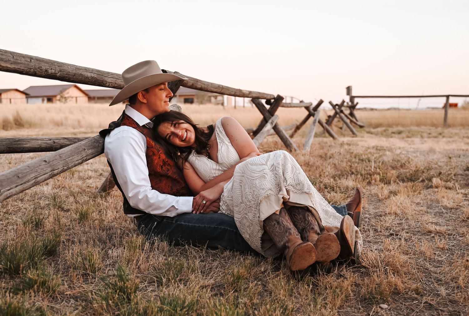 Couple in western attire relaxing against wooden fence in rustic setting.