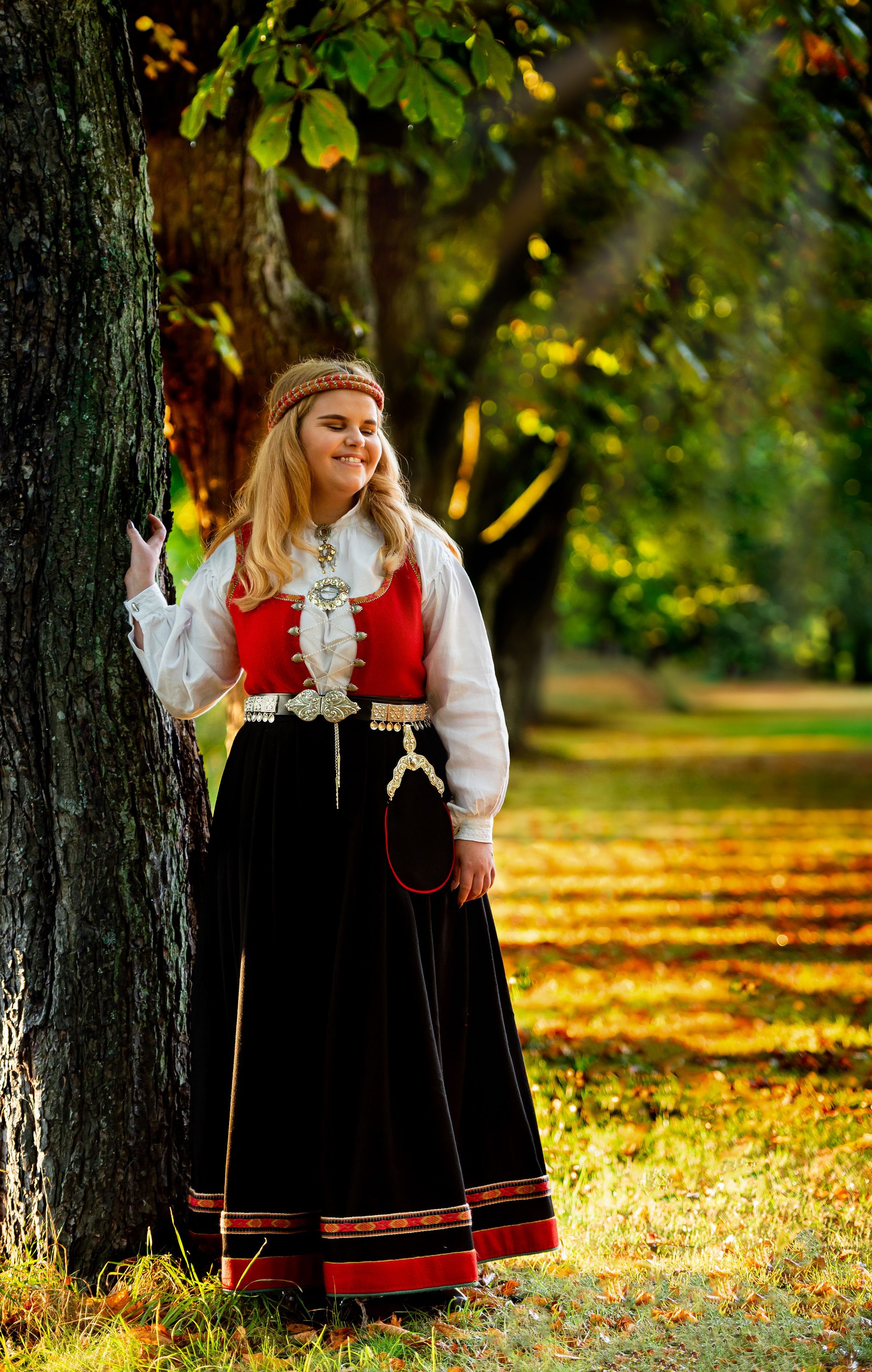 Woman in traditional Norwegian folk costume standing by tree in autumn park.