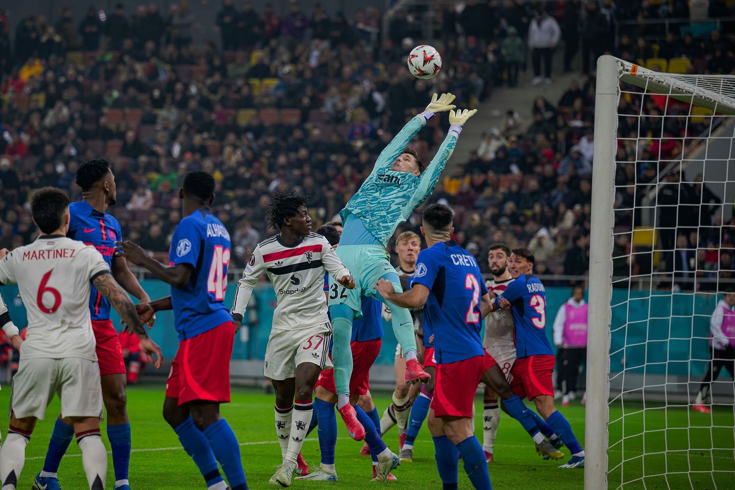 Soccer players from different teams leap for the ball as the goalkeeper reaches up to make a save during a match.