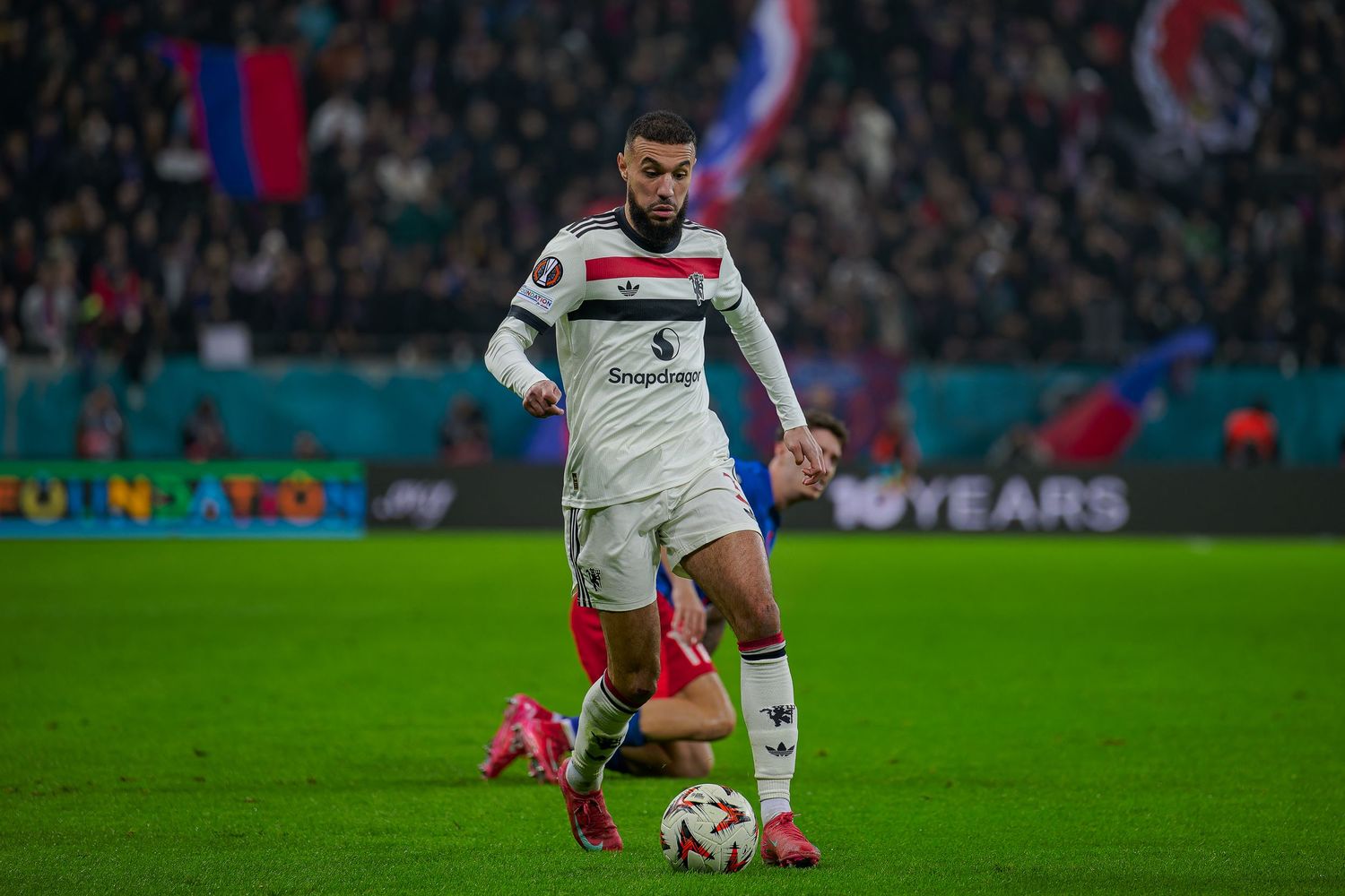 Soccer player in white jersey controls ball during match at crowded stadium under floodlights.
