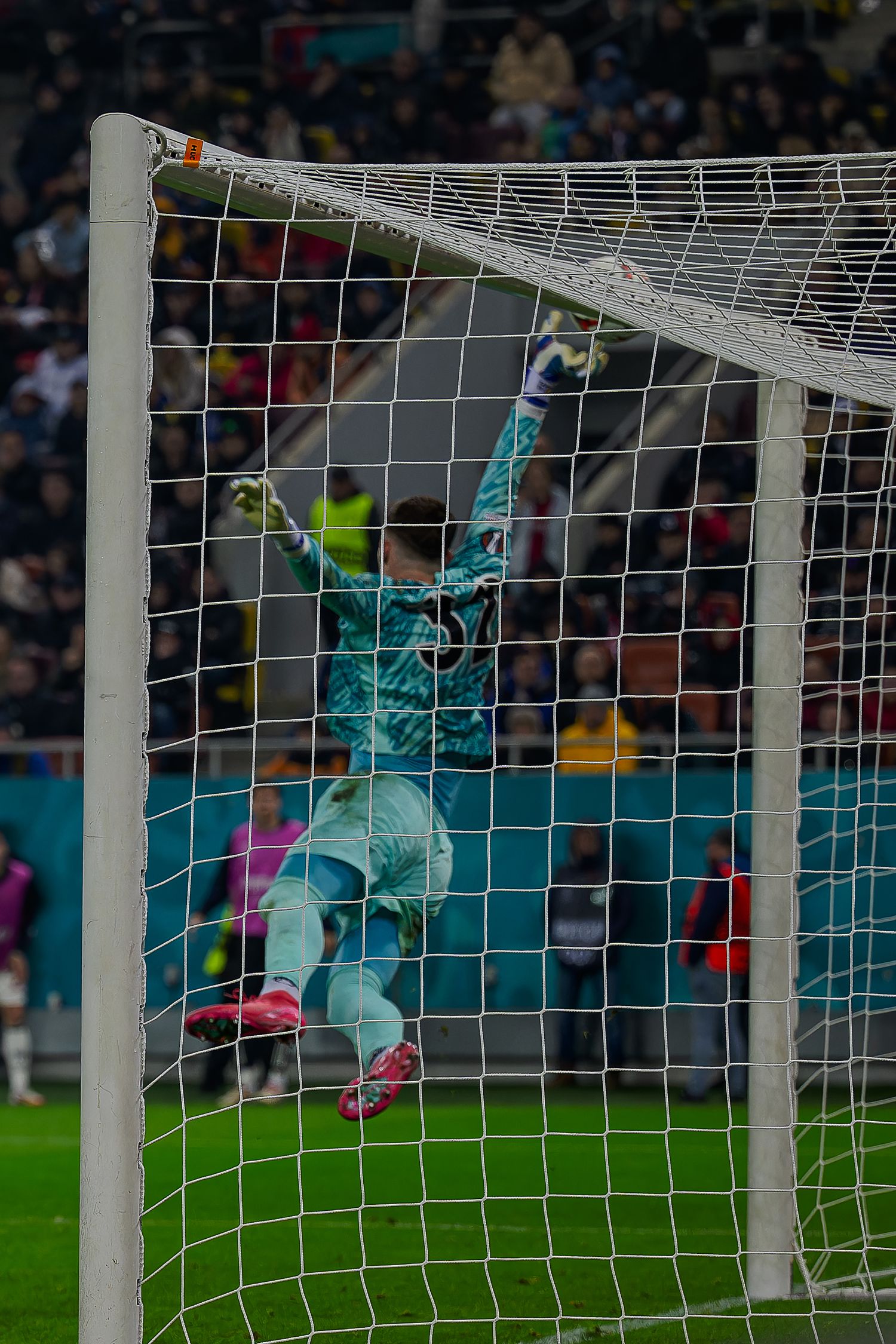 A soccer goalkeeper in turquoise uniform makes a leaping save at the goalpost during a match at a stadium.