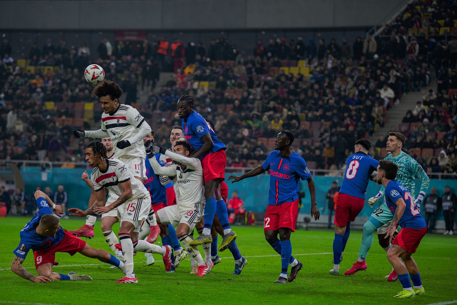 Soccer players from two teams leap for a header during a competitive match in a crowded stadium under floodlights.