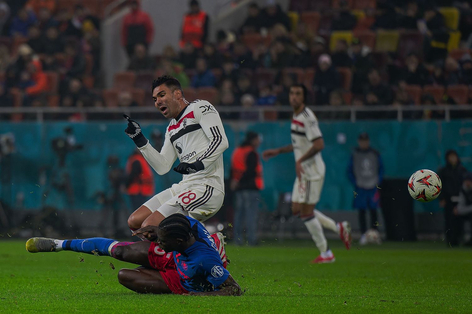 Soccer players compete for the ball during a match with a sliding tackle on a wet field under stadium lights.