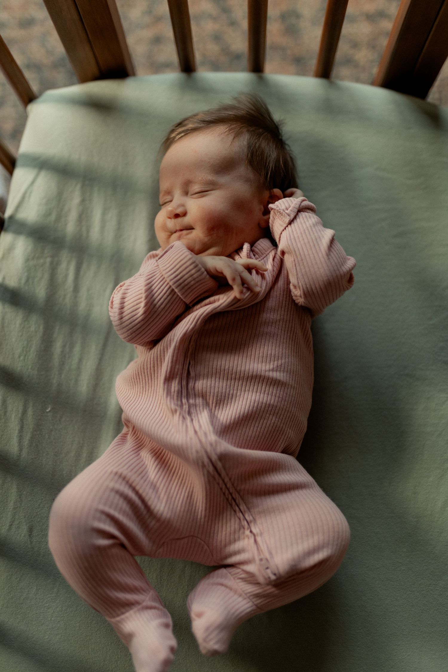 Sleeping baby wearing pink ribbed pajamas rests peacefully in a wooden crib.