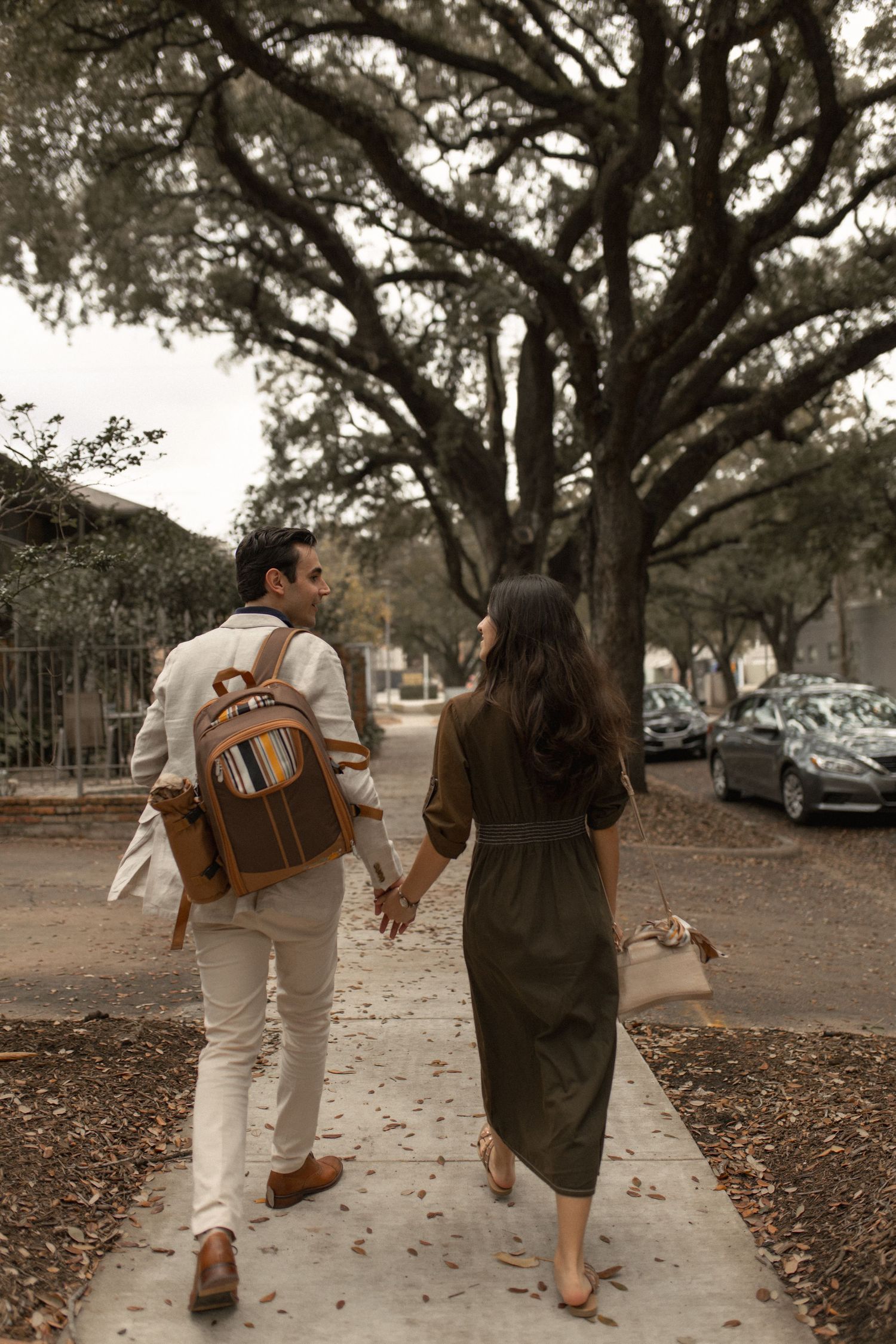 Couple walking hand in hand down a tree-lined sidewalk carrying a leather backpack.