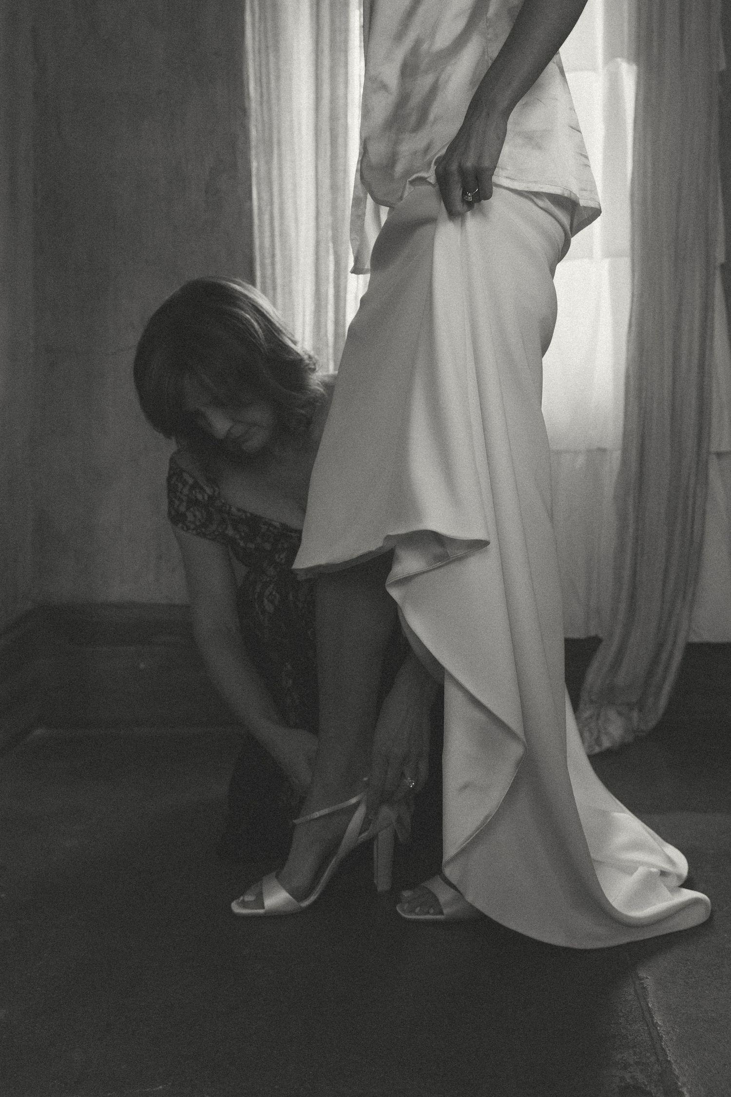 Black and white photo of someone adjusting a flowing wedding dress hem near a window.