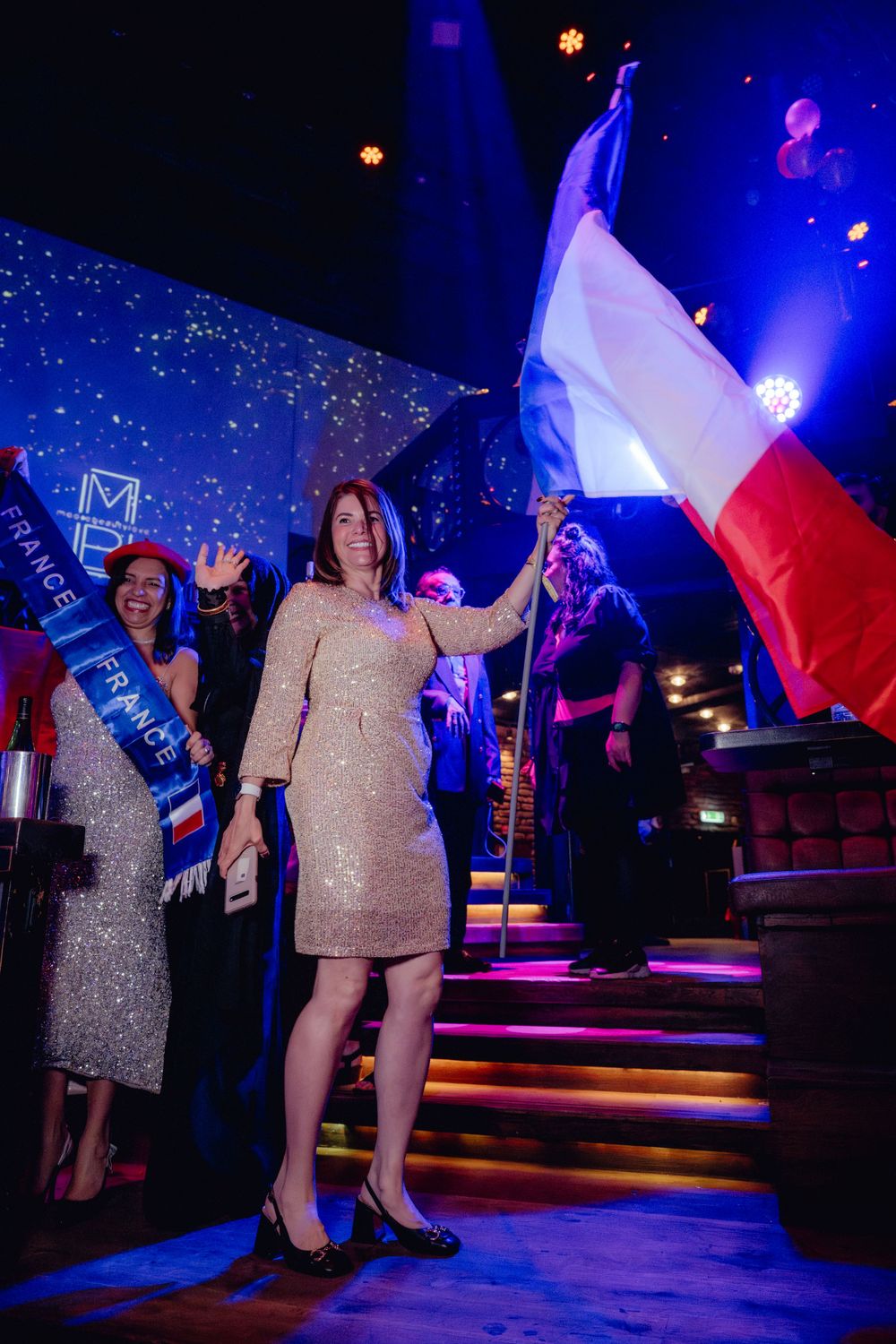 Person in sparkly dress holding French flag on illuminated stage steps with blue lighting.